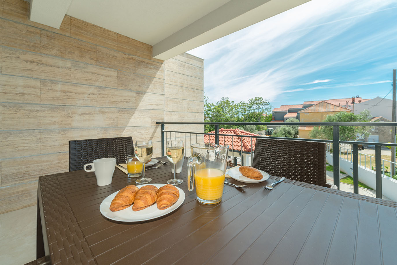 Sunčani balkon apartmana Vir s postavljenim stolom za doručak, s kroasanima, sokom od naranče i vinom, te slikovitim pogledom na krovove i zelenilo