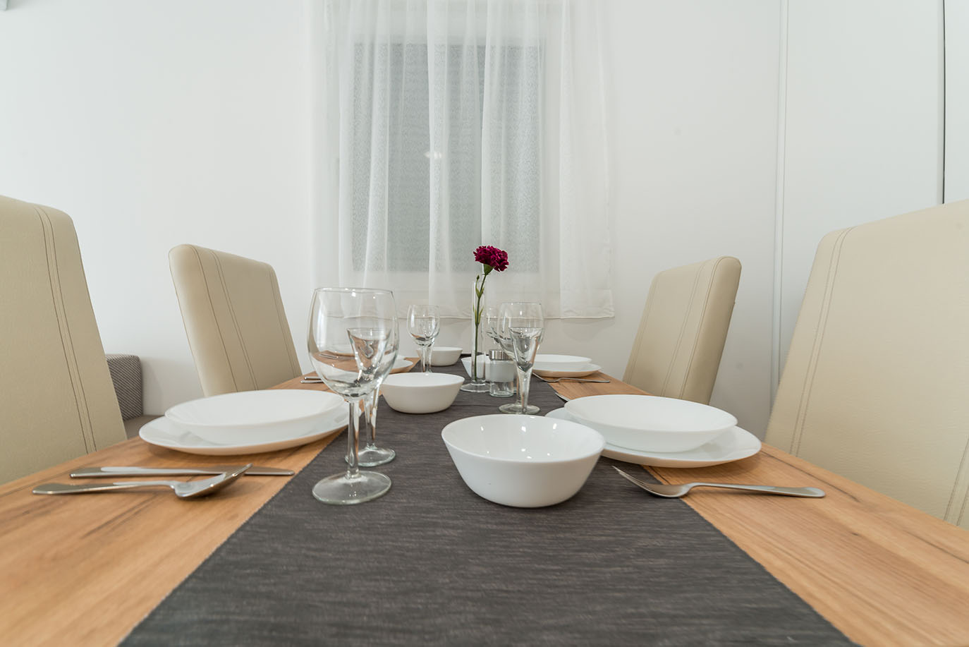 Svijetla blagovaonica s drvenim stolom postavljenim bijelim tanjurima, zdjelama, čašama za vino i priborom za jelo na tamnosivom nadstolnjaku, spremna za goste u kvalitetnim apartmani Vir