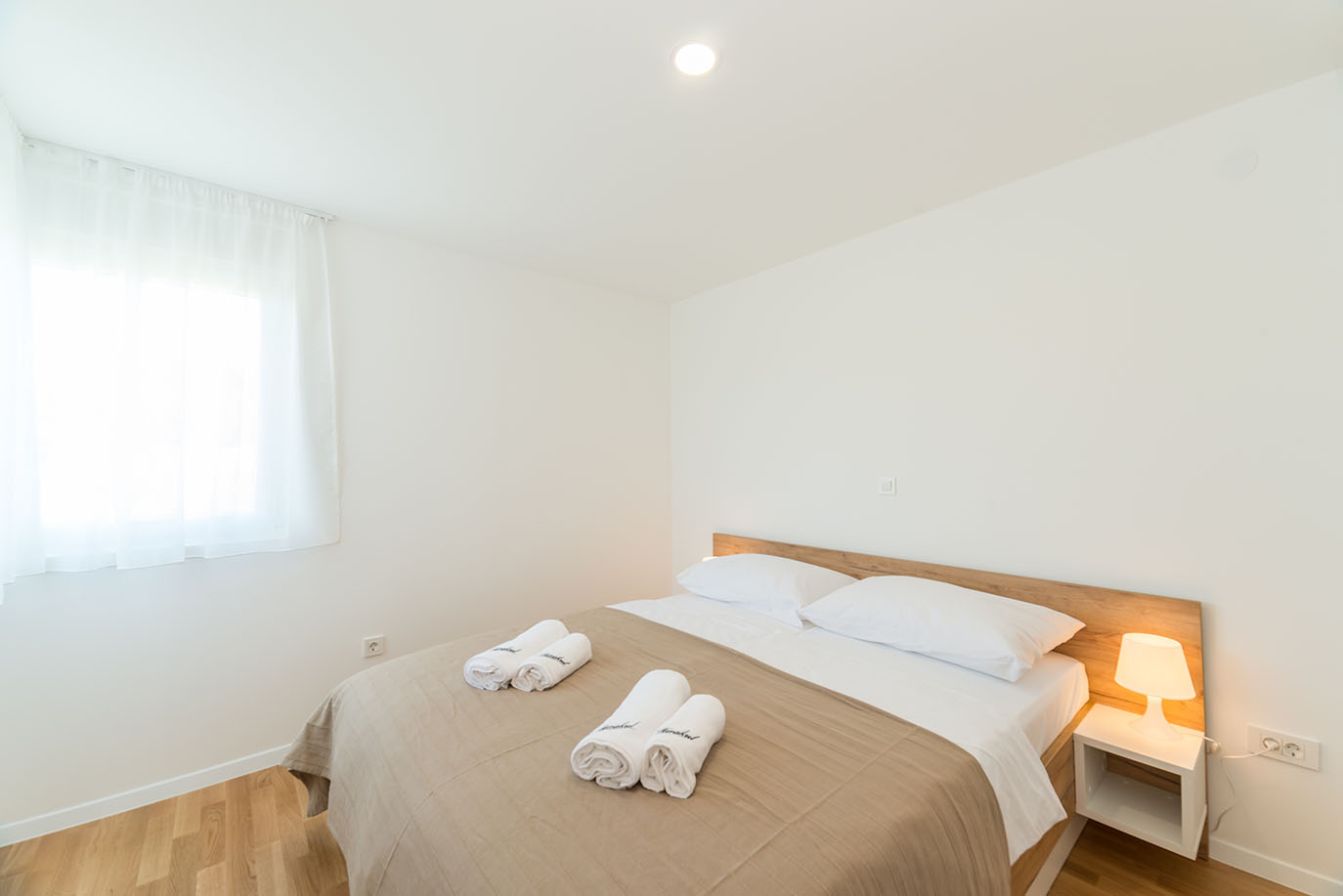 Svijetla i moderna dvokrevetna spavaća soba s drvenim uzglavljem, besprijekornom bijelom posteljinom i dvama setovima smotanih ručnika, idealna za goste koji odsjedaju u apartmanima Vir