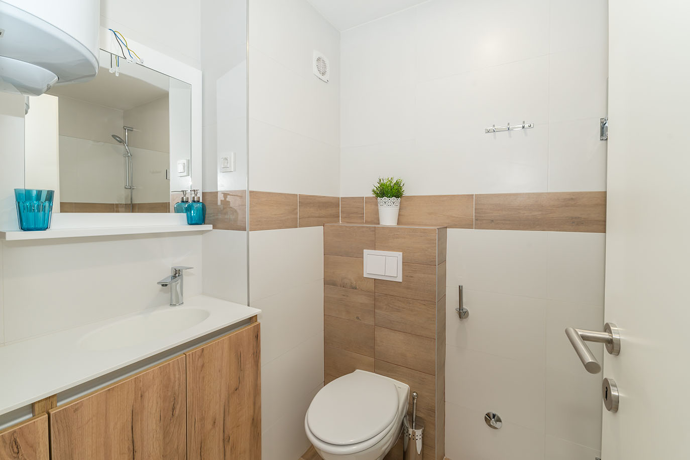 Moderna kupaonica u Villi Mirakul, opremljena elegantnim drvenim ormarićem, integriranim umivaonikom, velikim ogledalom s policom i čistim WC-om, nadopunjena bijelim pločicama i pločicama imitacije drva