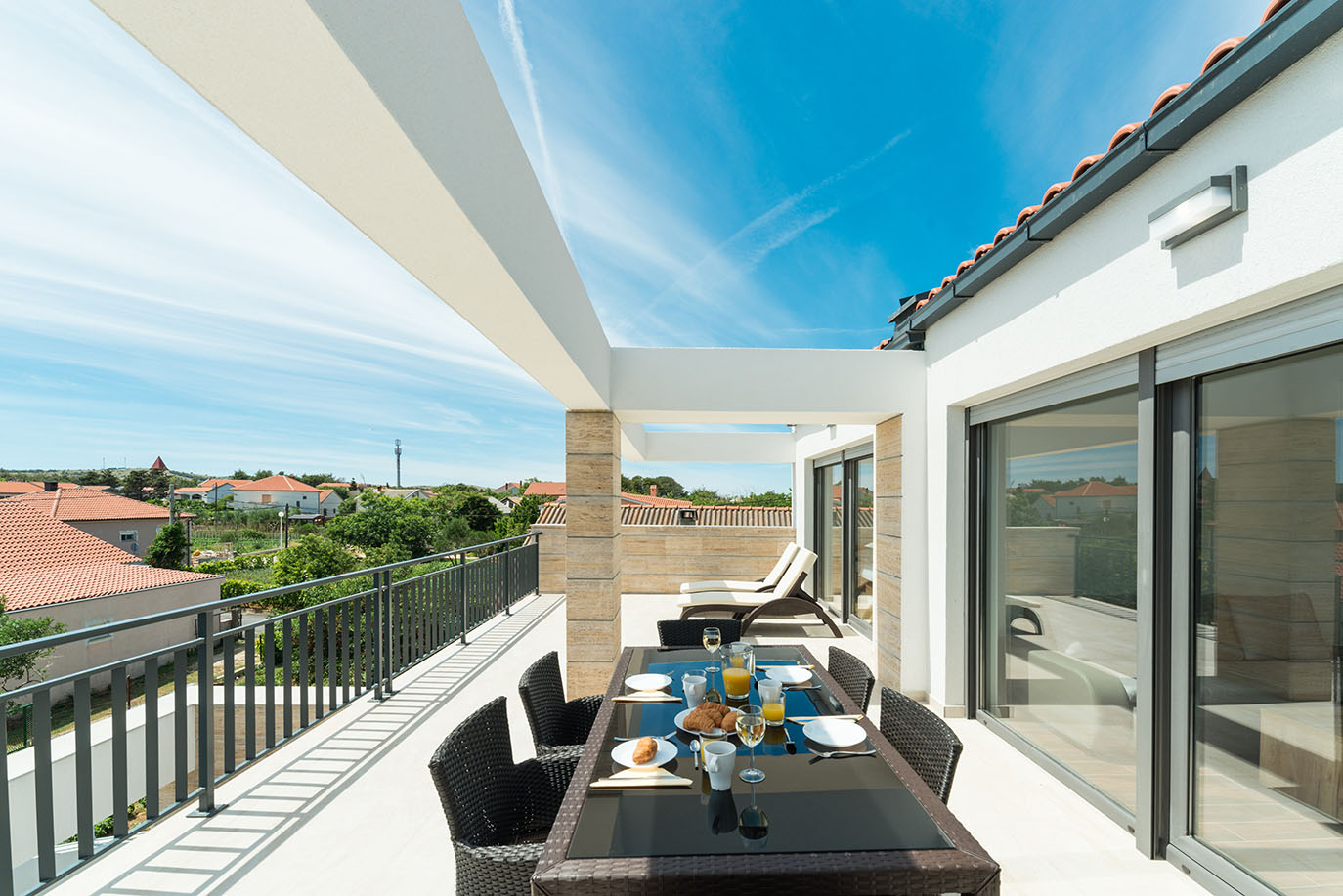 Prostrana, sunčana terasa Ville Mirakul s postavljenim stolom za doručak za četiri osobe, ležaljkama i panoramskim pogledom na hrvatsko selo pod vedrim plavim nebom