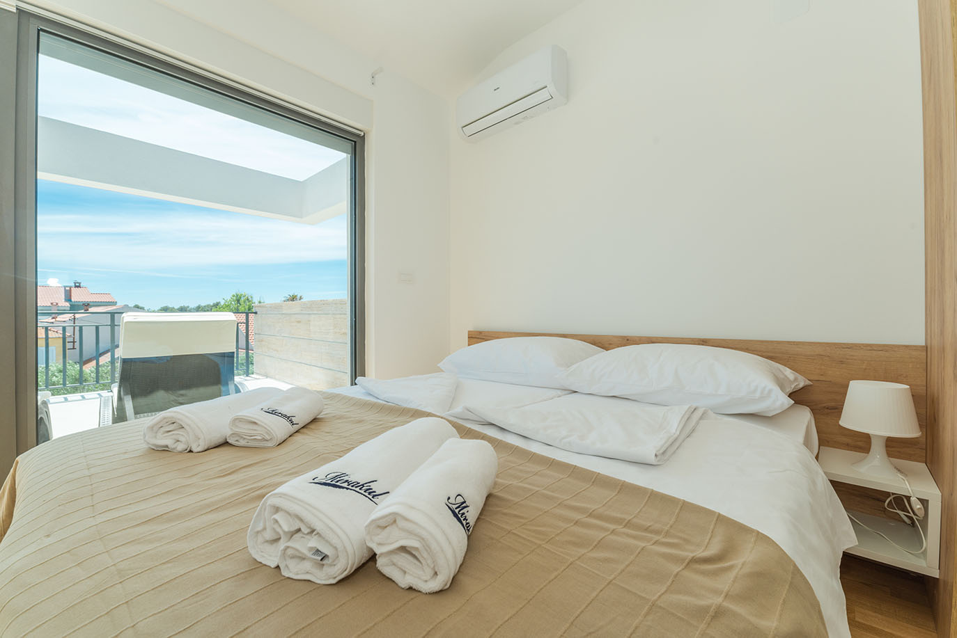Svijetla spavaća soba u Villi Mirakul s udobnim krevetom, bijelim ručnicima s izvezenim natpisom 'Mirakul' i velikim staklenim vratima koja se otvaraju na balkon