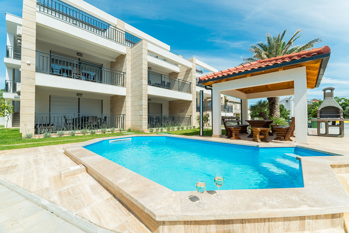 Moderna zgrada s balkonima, blistavim plavim bazenom, natkrivenim vanjskim prostorom za blagovanje i kamenim roštiljem, idealna za goste koji traže apartmane Vir