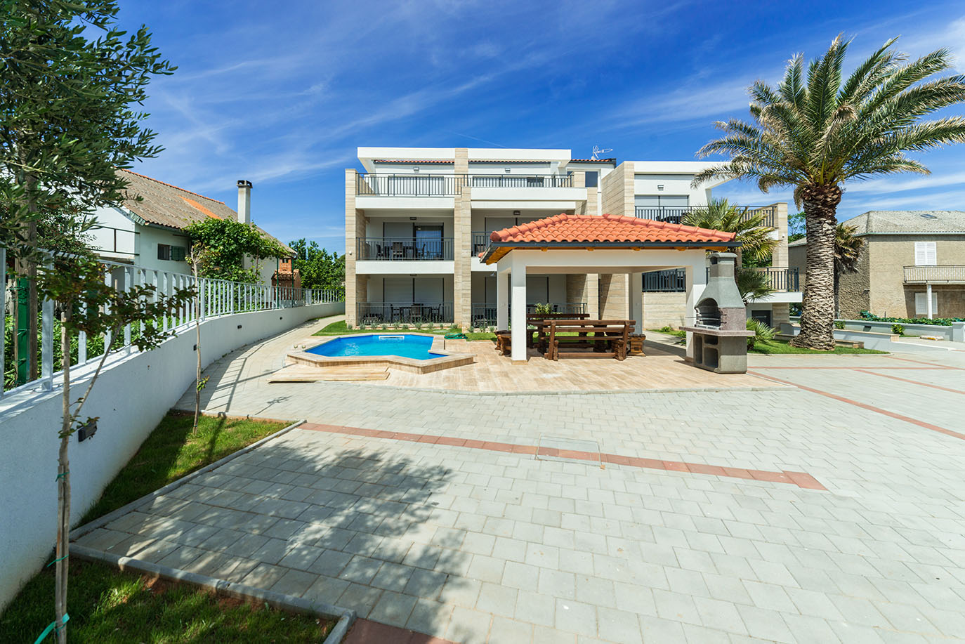 Vanjski snimak moderne Ville Mirakul, s blistavim plavim bazenom, natkrivenim prostorom za objedovanje s drvenim stolom i kamenim roštiljem za sunčanog dana