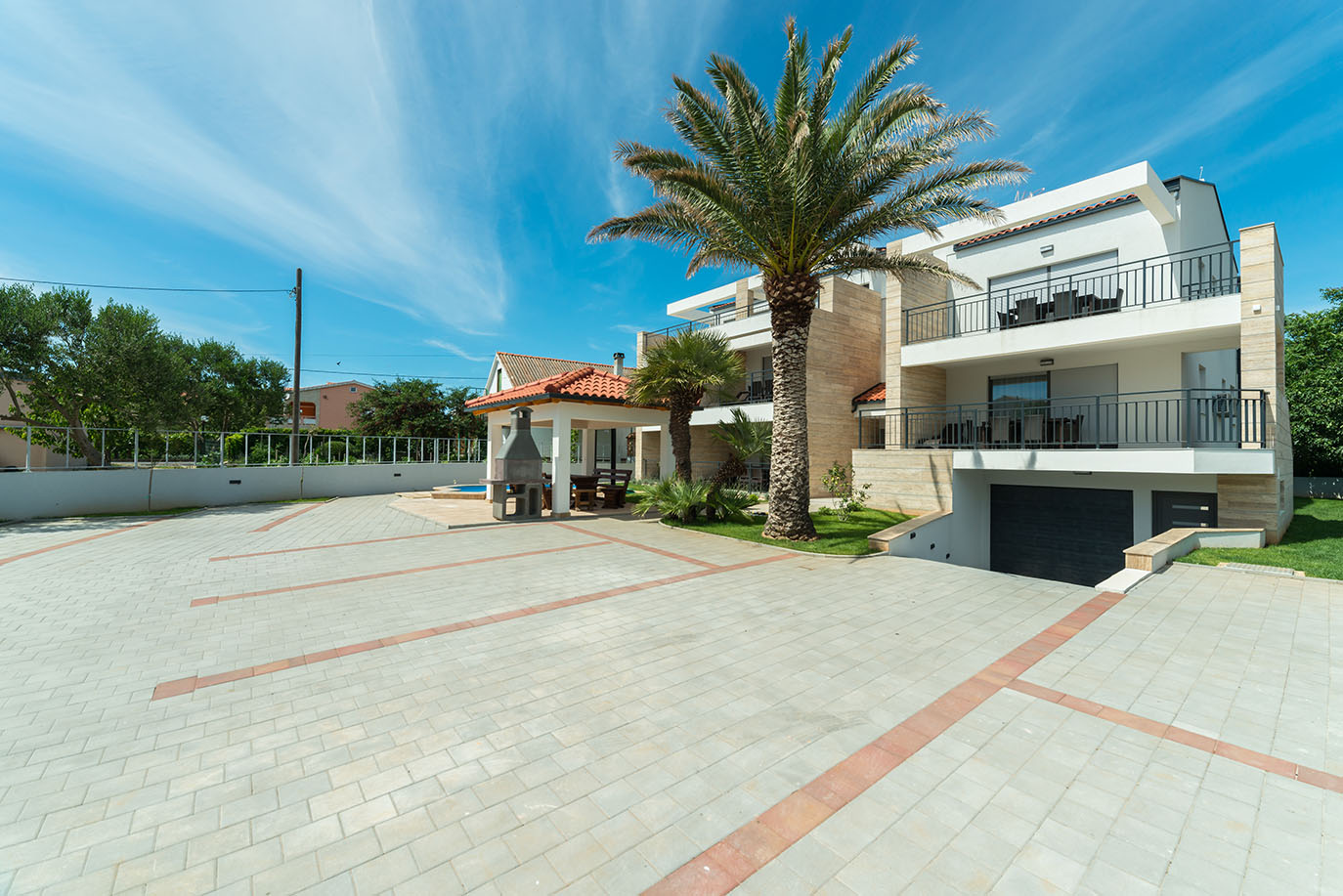 Vanjski izgled modernog objekta koji pruža luksuzni smještaj, s prostranim popločanim parkingom, zelenim palmama i privlačnim vanjskim roštiljem